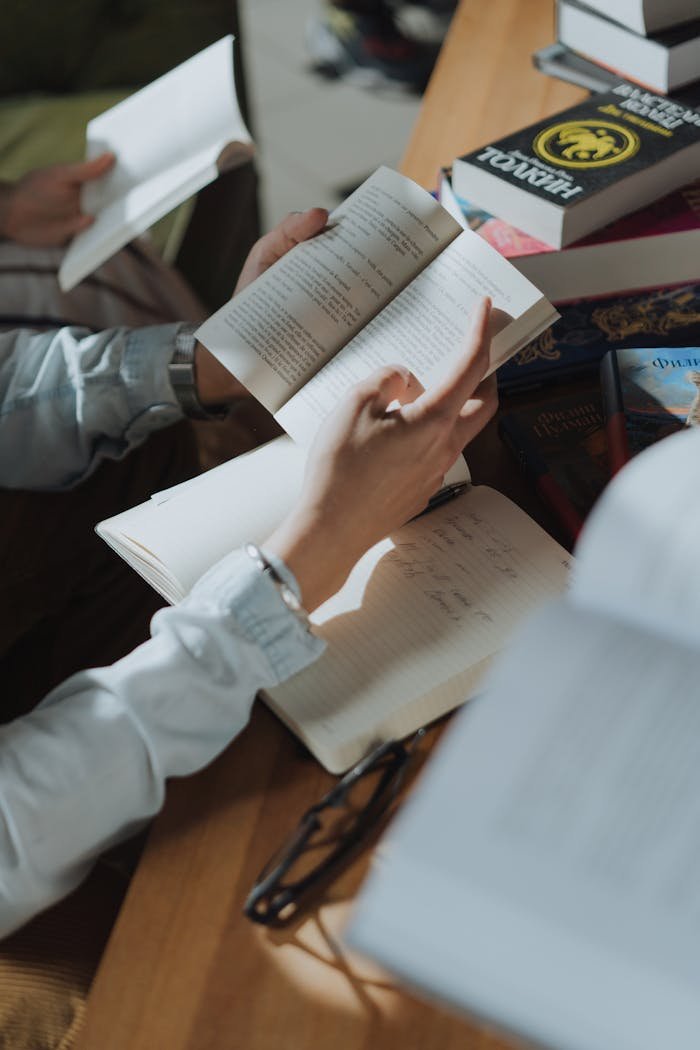 Person writing in a notebook while reading, surrounded by books in a cozy study setting.